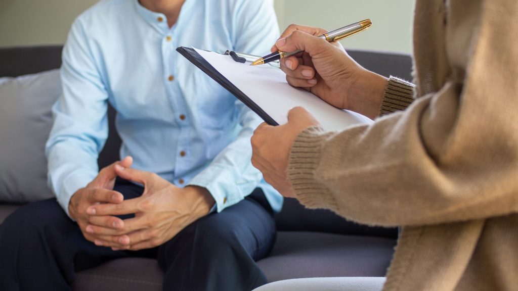 a man is sitting on a couch with a clipboard, discussing the importance of maintaining a healthy diet.