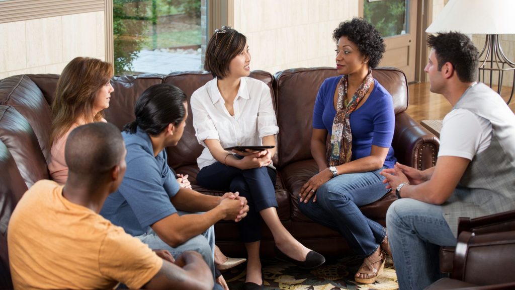 a group of six people, seated on a couch and chairs, engaged in discussion about living with hidradenitis suppurativa in a well-lit living room.