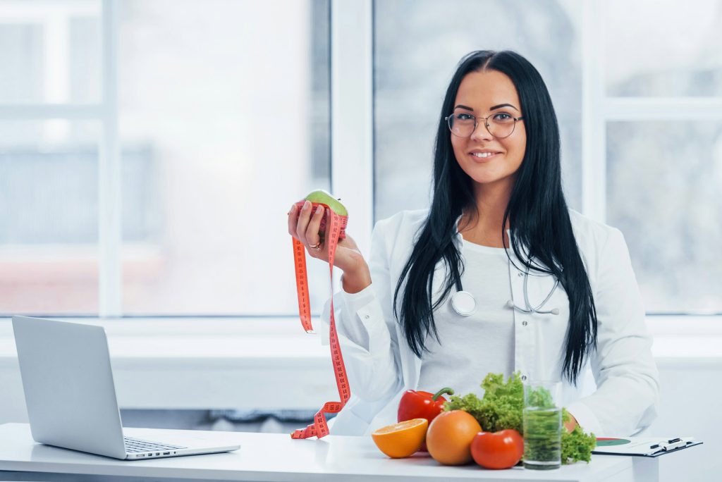 a woman in a lab coat holds an apple and a measuring tape while sitting at a table with fruits, her laptop open to research on diet.