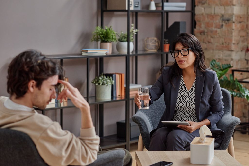 a person sits with their hand on their forehead, looking distressed, while another individual in glasses offers a glass of water in a room filled with bookshelves and plants, fostering an atmosphere focused on emotional well-being.