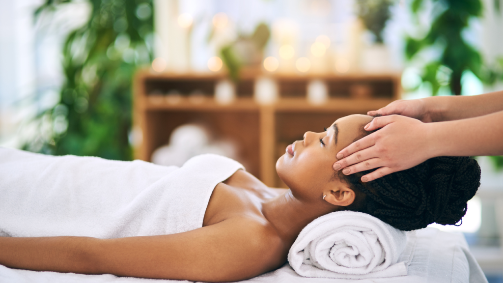 a woman lies on a massage table with a white towel, receiving a head massage in a tranquil setting with plants and blurred lights in the background, finding relief through pain management in hidradenitis suppurativa.