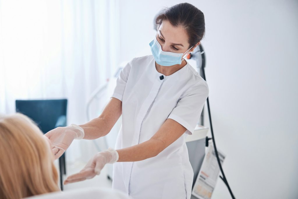 a medical professional wearing a mask and gloves examines a patient in a white room, preparing for laser therapy for hs.