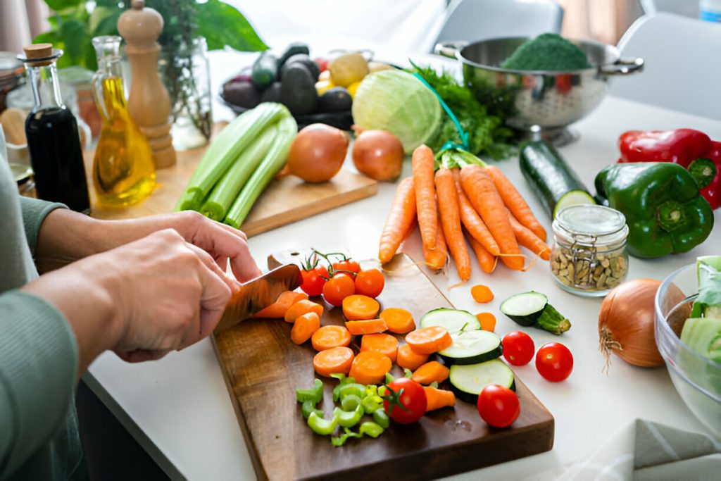 person chopping fresh vegetables, including carrots, celery, and tomatoes, on a wooden cutting board. various other vegetables and ingredients are spread across the kitchen counter, showcasing a focus on diet and nutrition in managing hidradenitis suppurativa symptoms.