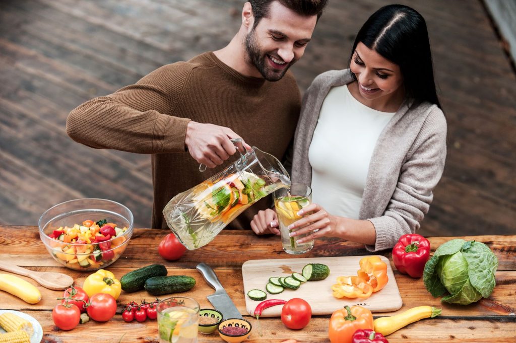 a man pours fruit-infused water from a jug into a woman's glass at a wooden table adorned with fresh vegetables and fruits, discussing pain management in hidradenitis suppurativa.