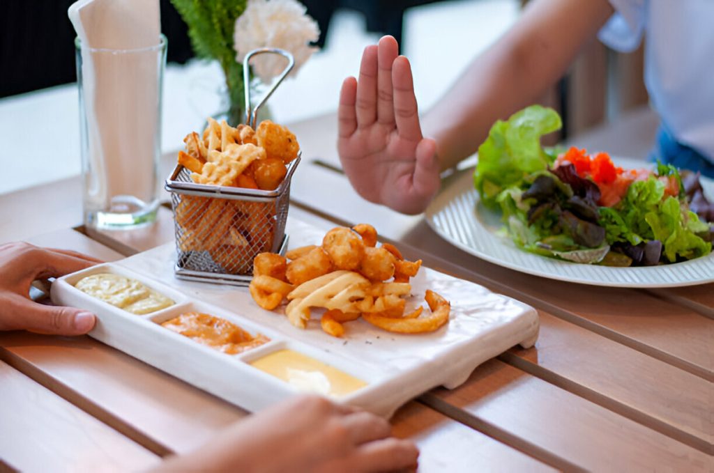 a person is holding up their hand, gesturing "stop" at a tray of fried food on a table while another person reaches toward it. a plate of salad is also visible on the table, underscoring the importance of diet and nutrition in managing hidradenitis suppurativa symptoms.