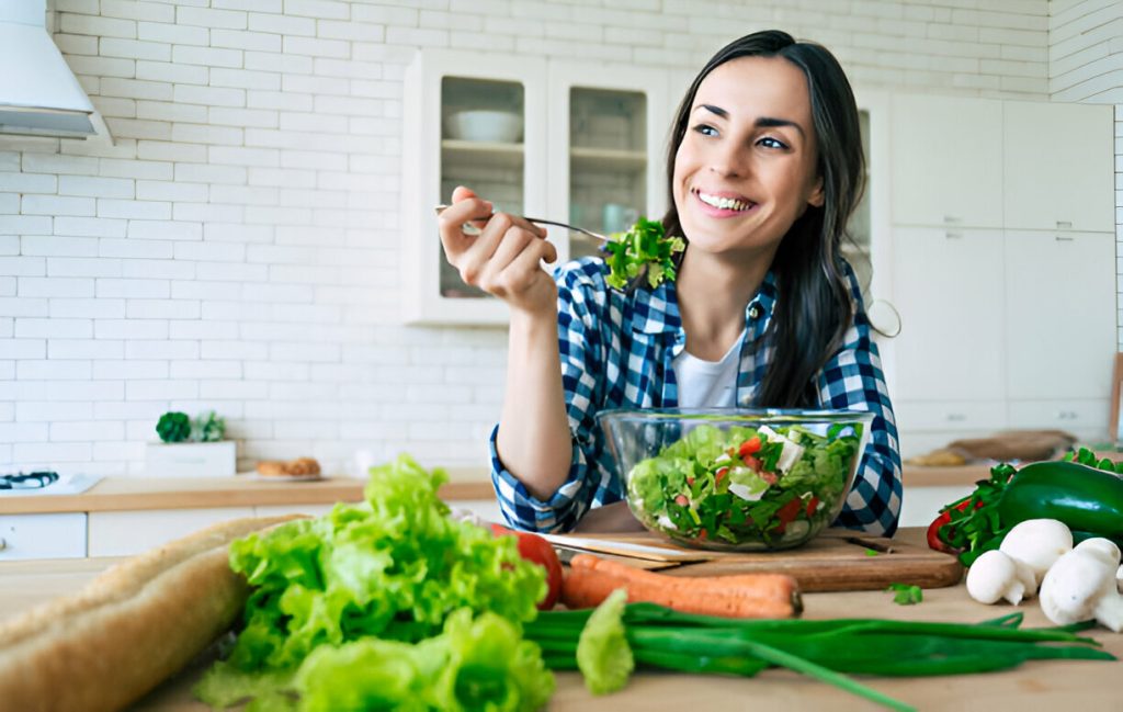 a woman in a checkered shirt is seated in a kitchen, smiling and eating a fresh salad from a bowl, surrounded by various vegetables on the counter, showcasing how diet and nutrition play a role in managing hidradenitis suppurativa symptoms.