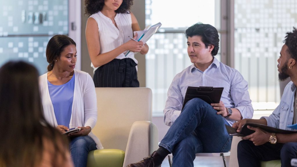 a group of people is engaged in a meeting; two seated individuals hold tablets, while a standing woman from the support network for hs patients holds a notepad and pen.