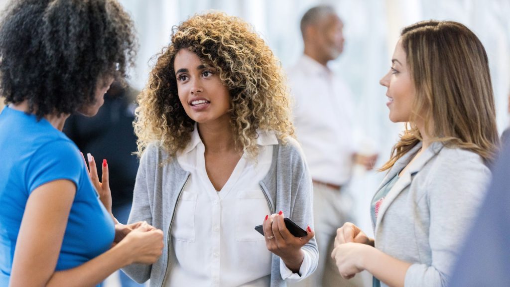 three women are engaged in a conversation at a social gathering. one woman holds a smartphone, sharing details about the support network for hs patients, while the other two listen attentively. other people are in the background.