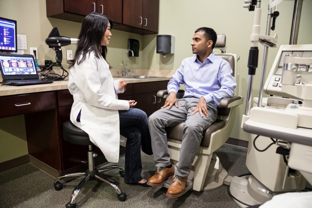 a healthcare professional in a white coat is sitting and talking with a patient in an examination room equipped with medical devices, a computer, and information on natural treatments.