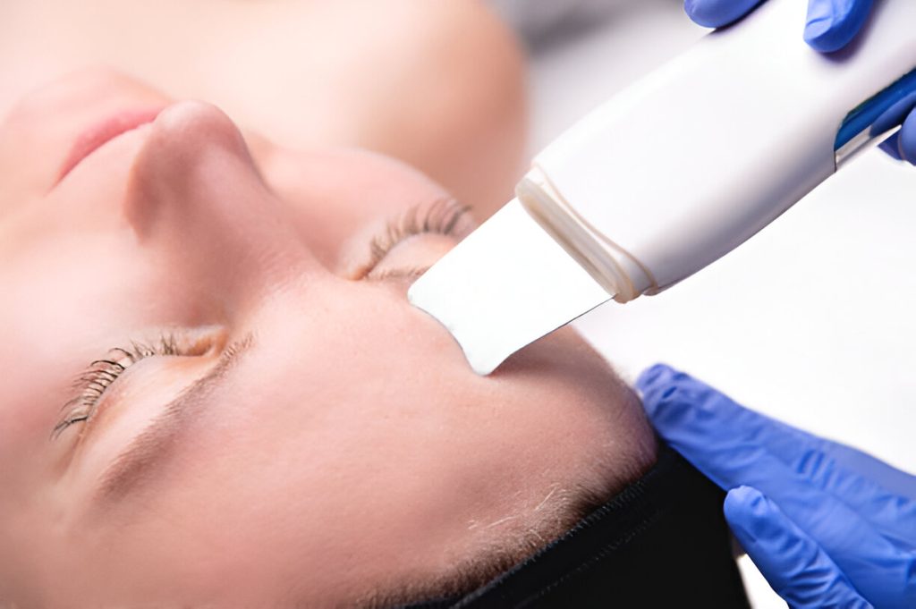 a person is receiving a beauty treatment with a white handheld device applied to their forehead by a practitioner wearing blue gloves, complimenting the topical treatments they have been using.