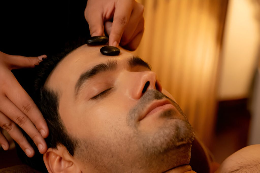 a person receiving a hot stone therapy massage, with one stone placed on their forehead and another about to be placed, is lying down with eyes closed, benefiting from these natural treatments.