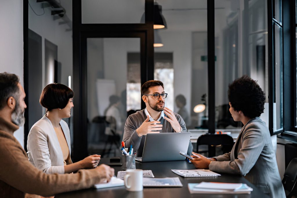 four people in a meeting room, one person speaking about managing hidradenitis suppurativa while others listen attentively around a table with laptops and papers.