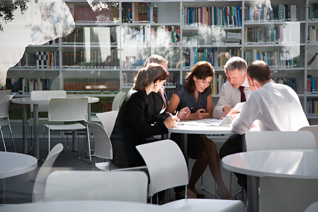 a group of five people sit around a table in a library, engaged in managing hidradenitis suppurativa through discussion. shelves lined with books are visible in the background.