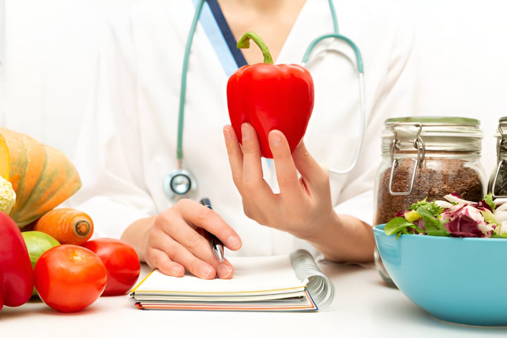 a person in a white coat with a stethoscope holds a red bell pepper while writing in a notebook, reflecting on natural treatments. various vegetables and a salad bowl are on the table.